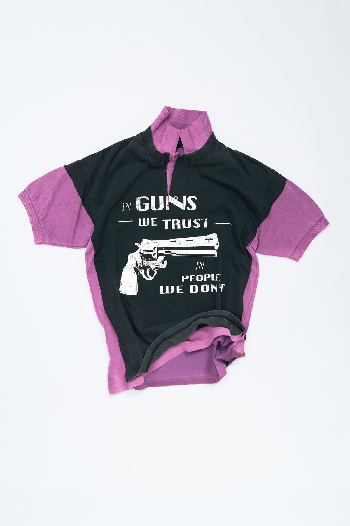 A short-sleeve polo shirt with a vintage black body and purple sleeves/collar, featuring a white graphic print of a revolver and the text 'IN GUNS WE TRUST' and 'IN PEOPLE WE DON'T'.