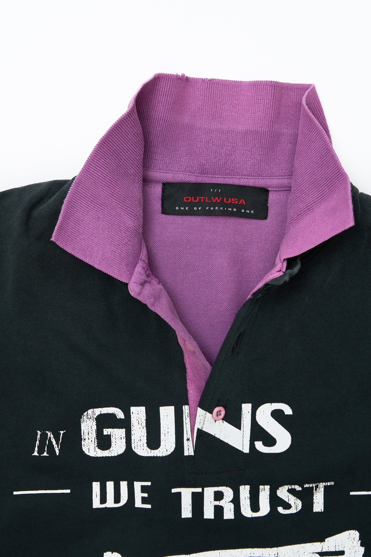 Close-up of a collared shirt with a purple inner collar lining and a visible neck label reading 'OUTLW USA'. The front has large white text reading 'IN GUNS WE TRUST'.