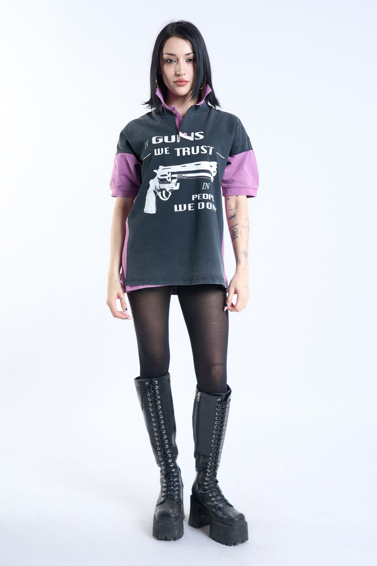 A model stands against a white studio background wearing an oversized dark gray graphic T-shirt with pink/purple sleeves and collar trim, black tights, and tall black lace-up boots. The shirt features a white revolver graphic and the text 'IN GUNS WE TRUST' and 'IN PEOPLE WE DON'T'.