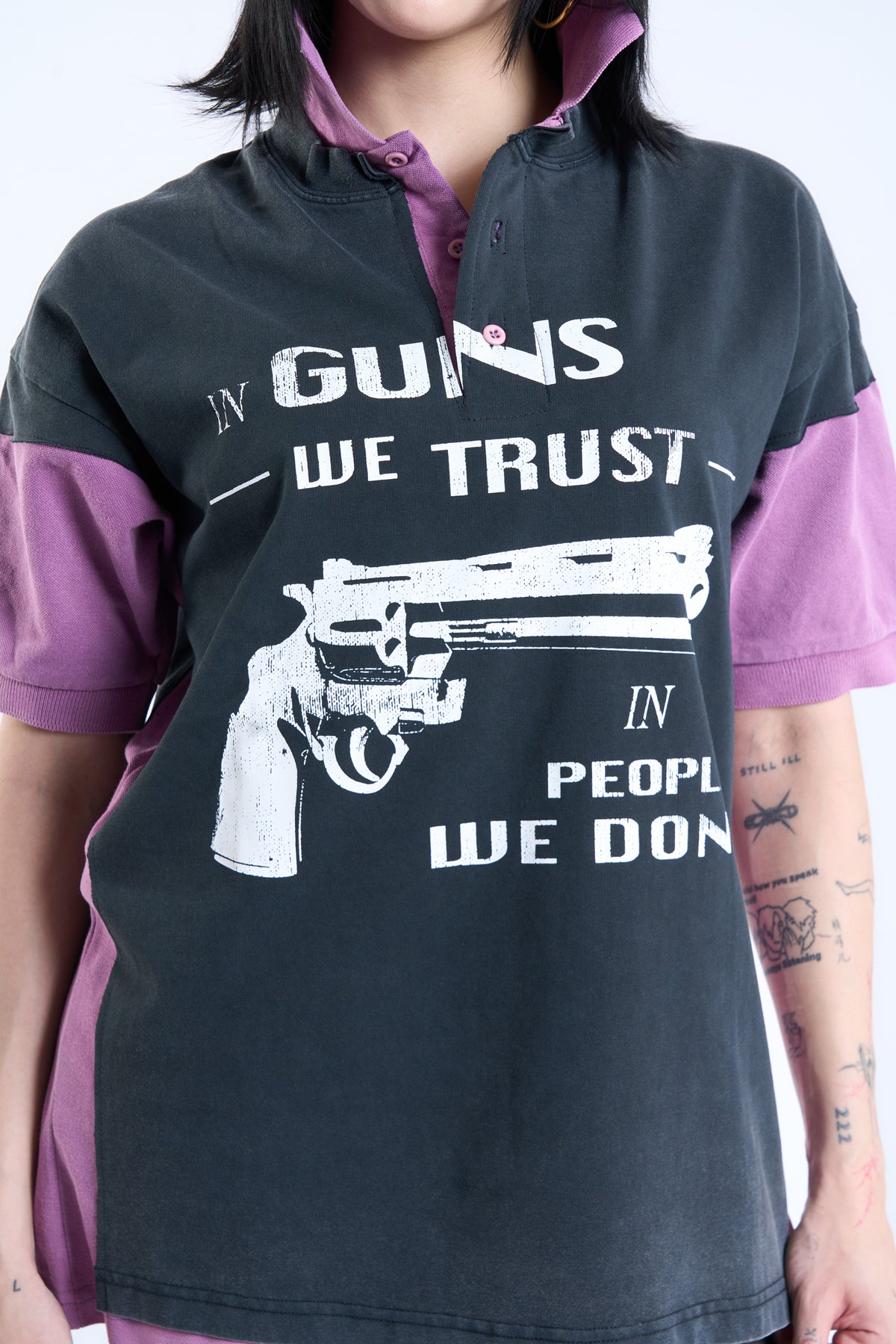 A person wearing a dark charcoal gray short-sleeve polo shirt with purple color-blocked sleeves and collar, featuring a white graphic of a revolver and the text 'GUNS WE TRUST' and 'PEOPLE WE DON'T'.