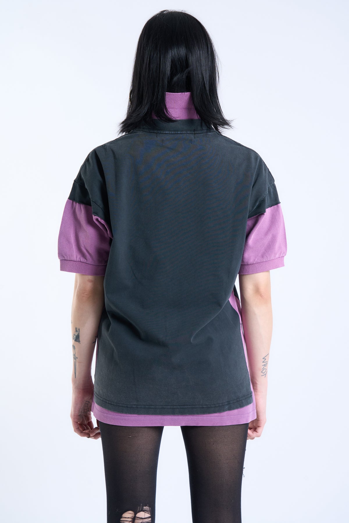 A model shown from the back wearing a short-sleeve polo shirt with a washed charcoal gray body and contrasting purple sleeves and hem, paired with black tights.